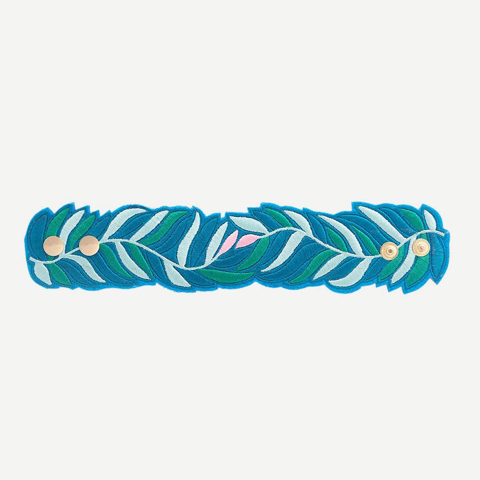 OMY besticktes Armband mit blauem, grünem und rosa Blattmuster, flach ausgelegt mit goldenen Druckknöpfen, auf weißem Hintergrund