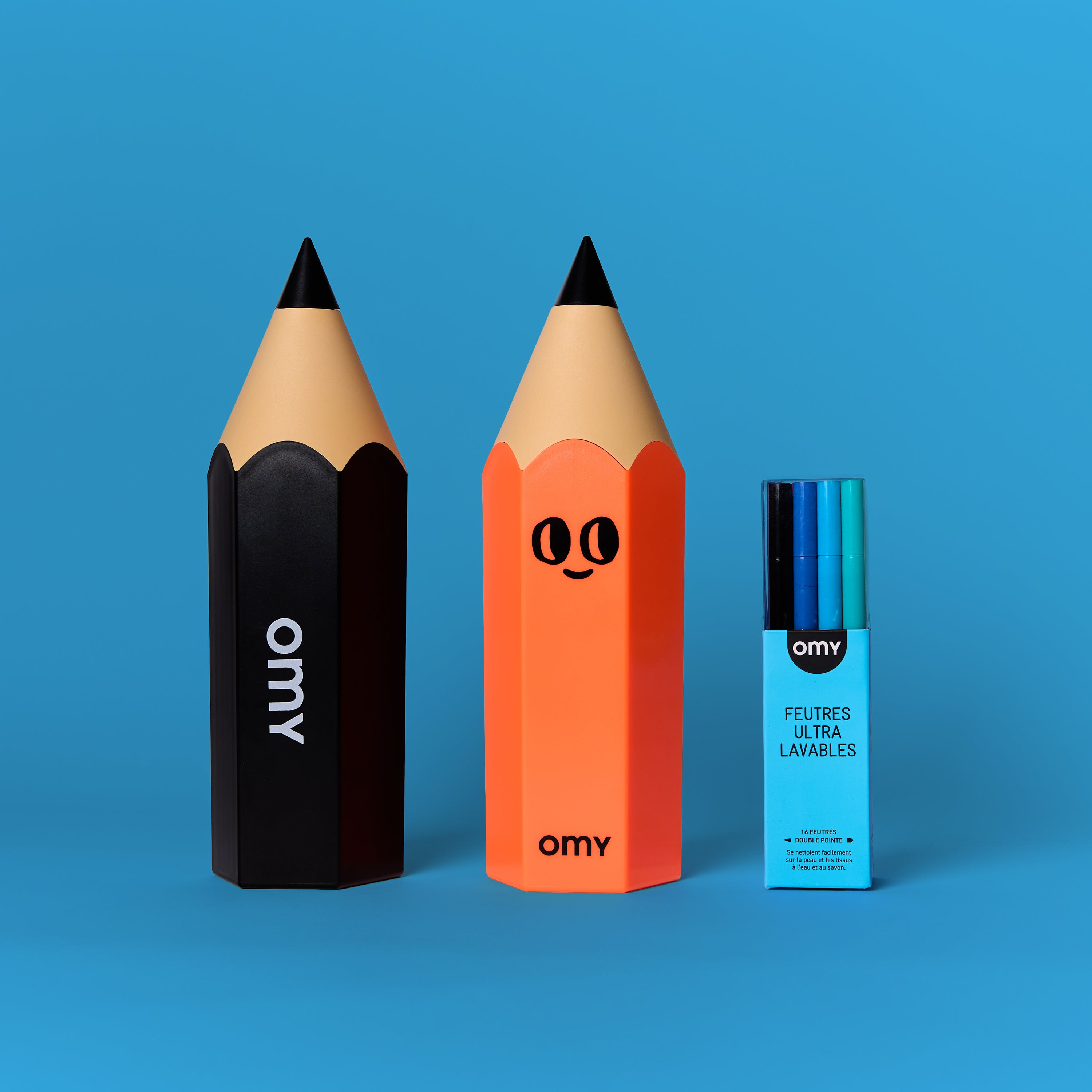 Deux boîtes à crayons géantes en forme de crayon OMY (noir et orange), accompagnées d’un étui de feutres ultralavables sur fond bleu vif.