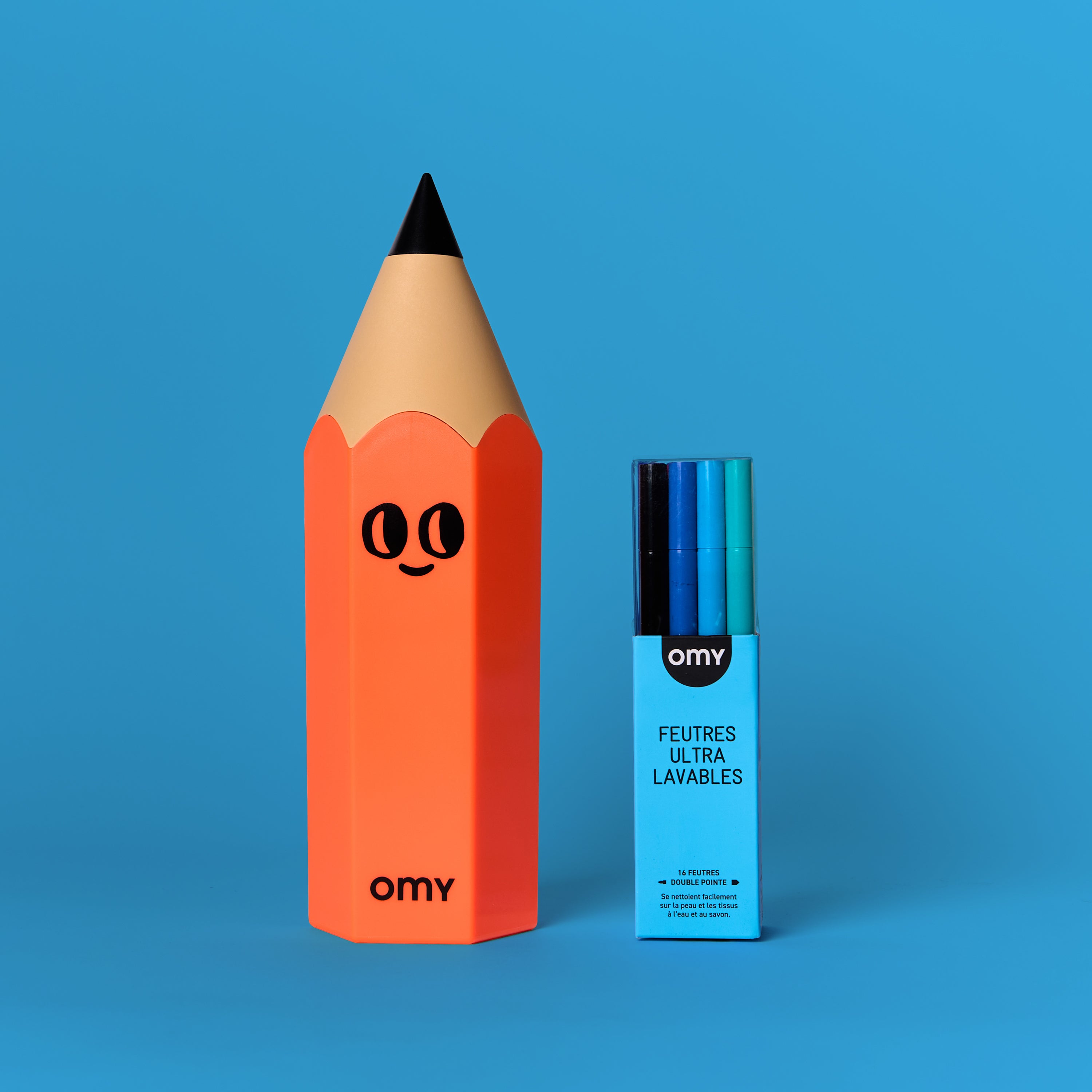 Boîte à crayons orange en forme de crayon avec un visage souriant et étui de feutres ultralavables bleu OMY, positionnés sur un fond uni bleu.