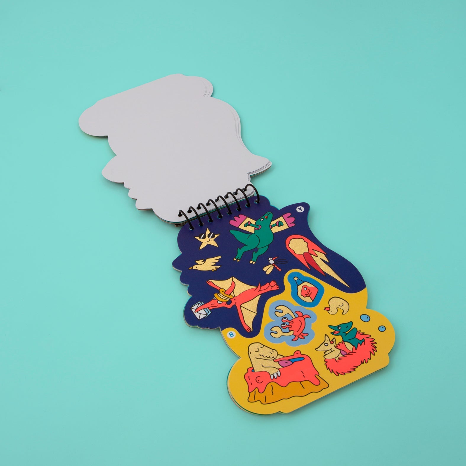 Carnet d'activités stickers Dino OMY ouvert sur une page de stickers dinosaures, météorite et fossiles, sur fond turquoise