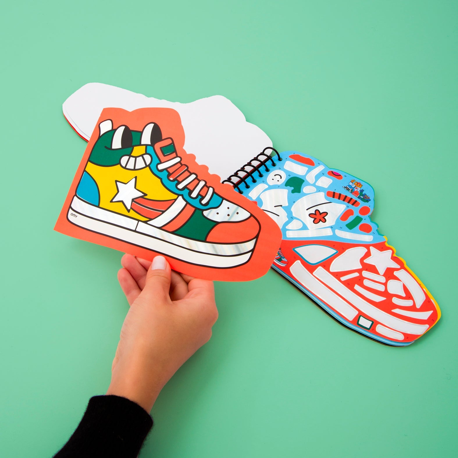 Carnet de stickers Street OMY en forme de basket, tenu en main, pages de stickers colorés ouvertes sur fond vert menthe