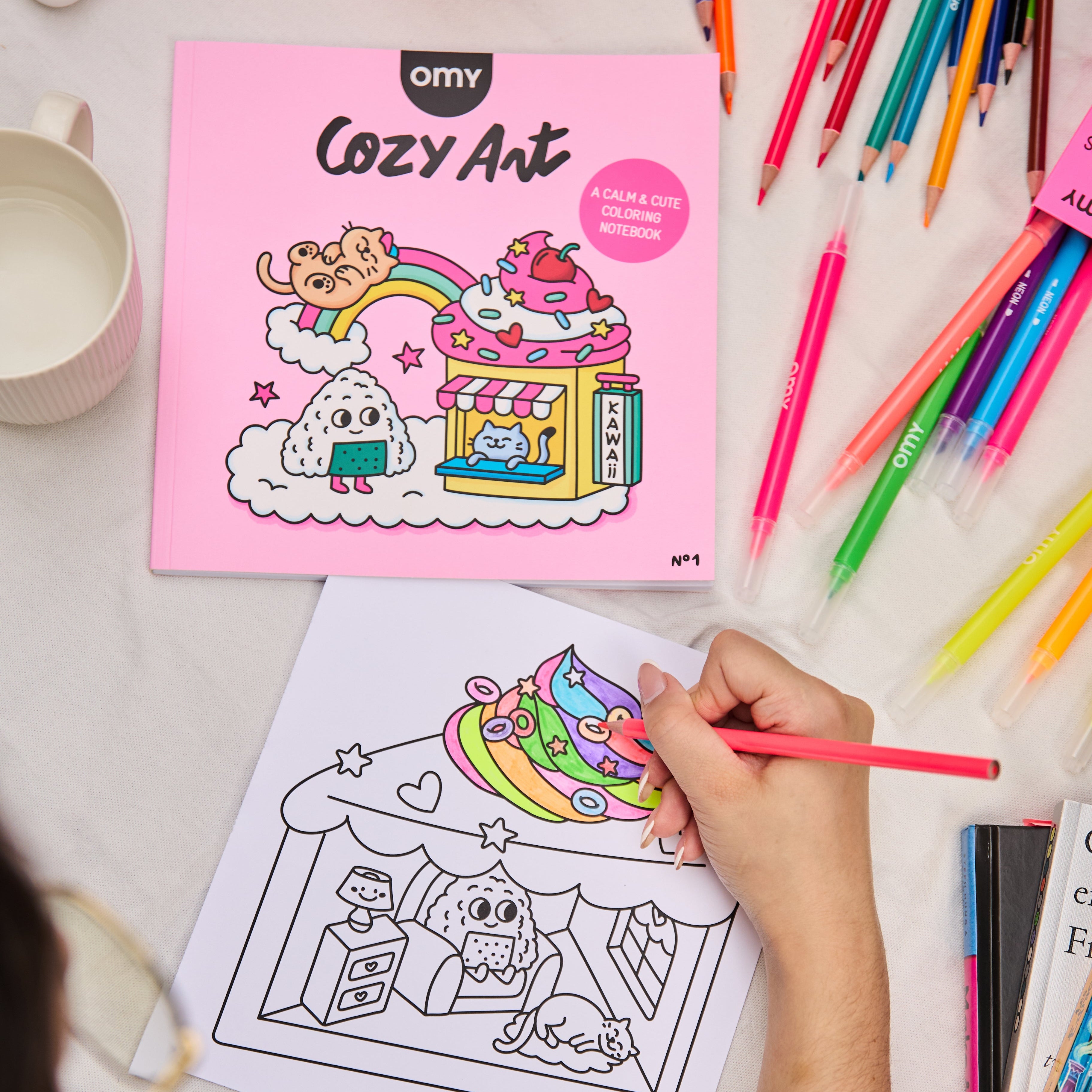 Enfant coloriant une illustration mignonne du carnet Cozy Art Kawaii d’OMY avec des feutres fluos, carnet posé à côté sur une table claire.