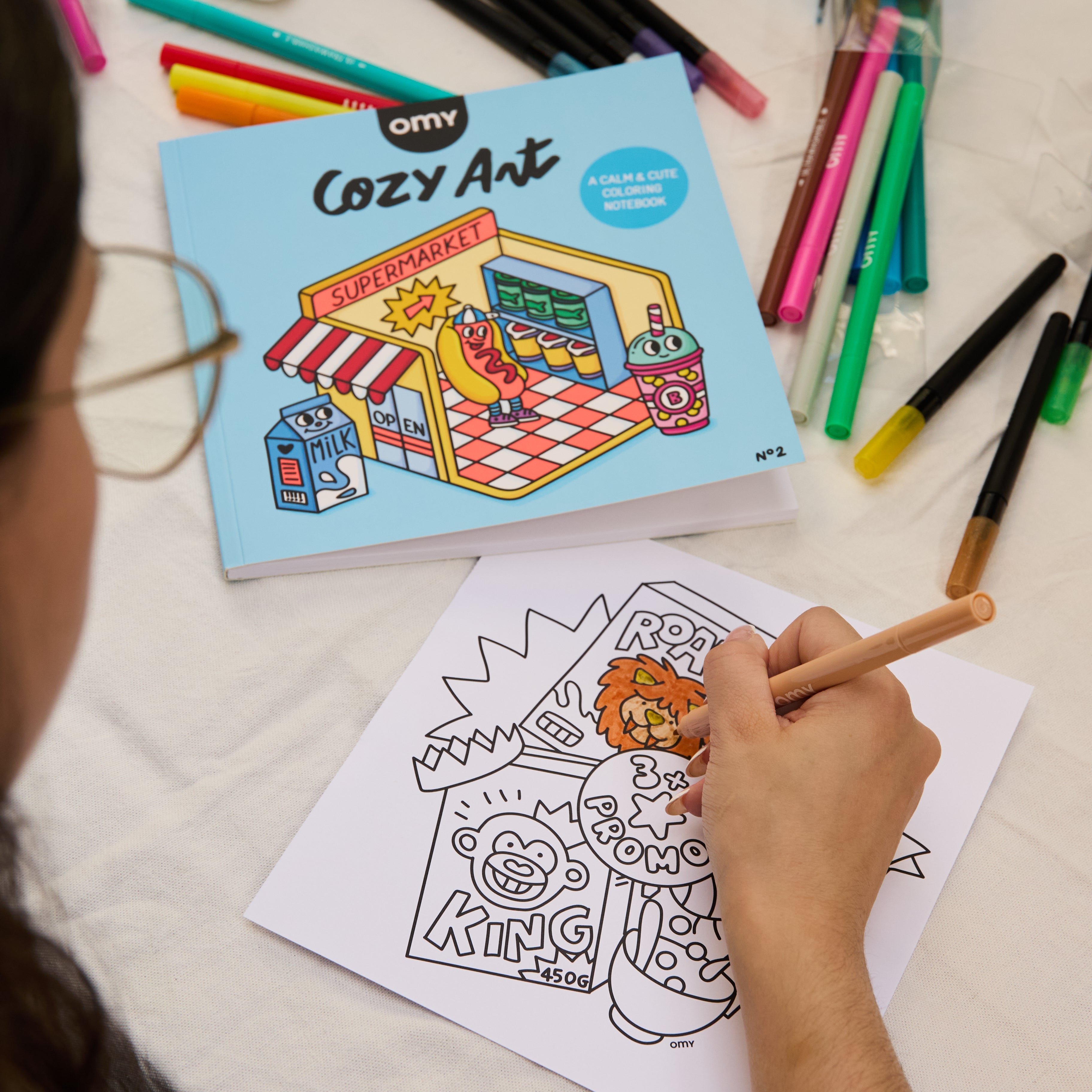 Enfant en train de colorier une illustration de lion issue du carnet Cozy Art Super Market d’OMY, carnet fermé en arrière-plan et feutres de coloriage étalés sur la table.