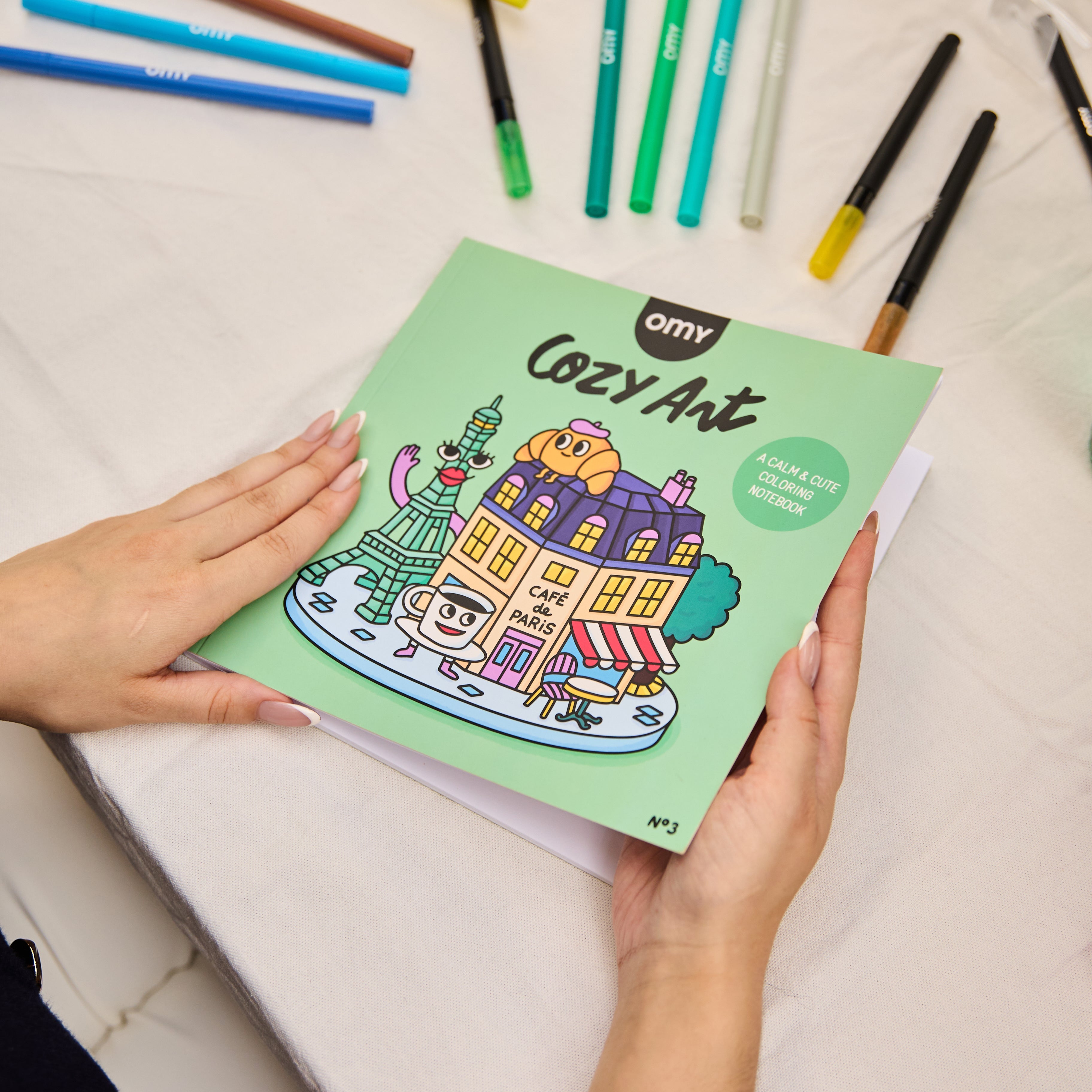 Main tenant le cahier de coloriage Cozy Art Paris d’OMY, posé sur une table blanche entourée de feutres, ambiance créative et apaisante.