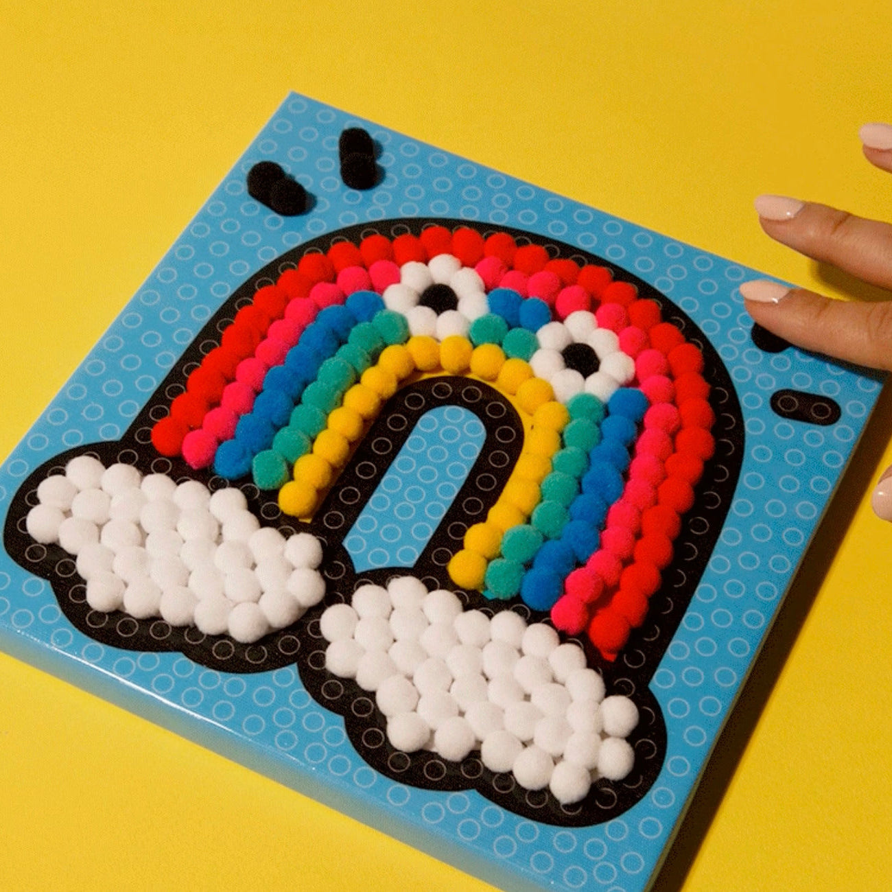 Main posant des pompons sur tableau Rainbow OMY – Développe créativité et motricité