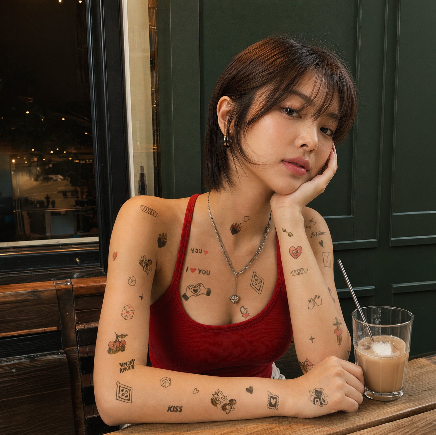 Flash tattoos temporaires OMY "Crazy In Love" portés par une jeune femme assise dans un café, avec des tatouages éphémères motifs amour, cœurs, "I Love You", "Kiss", "Je t'aime" et "Amour" sur les bras, la poitrine et les épaules.