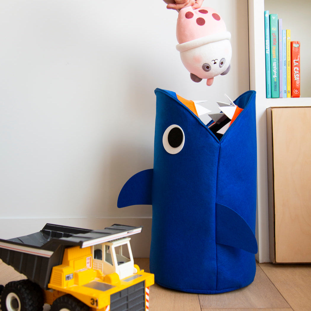 Tidy Bag Sharky OMY, blauer Aufbewahrungskorb für Kinder aus Filz in Form eines Hais, mit Kuscheltier und Laster in einem Kinderzimmer.