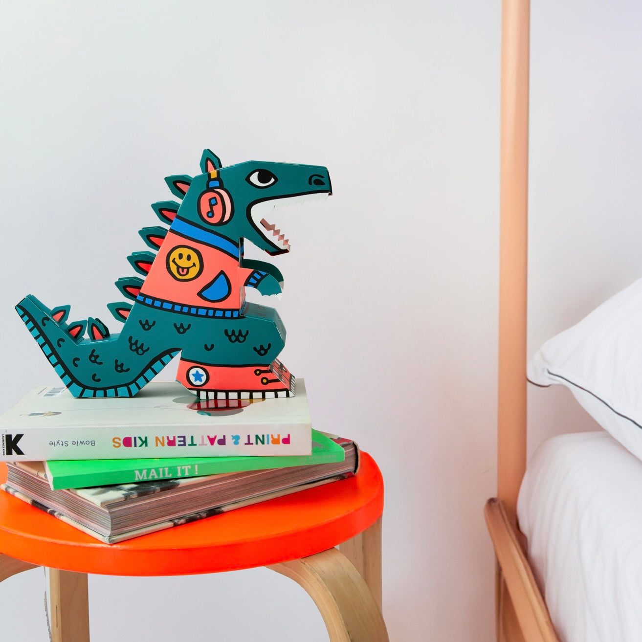 Lampe 3D OMY dinosaure vert et corail à casque audio, posée sur des livres et un tabouret orange dans une chambre