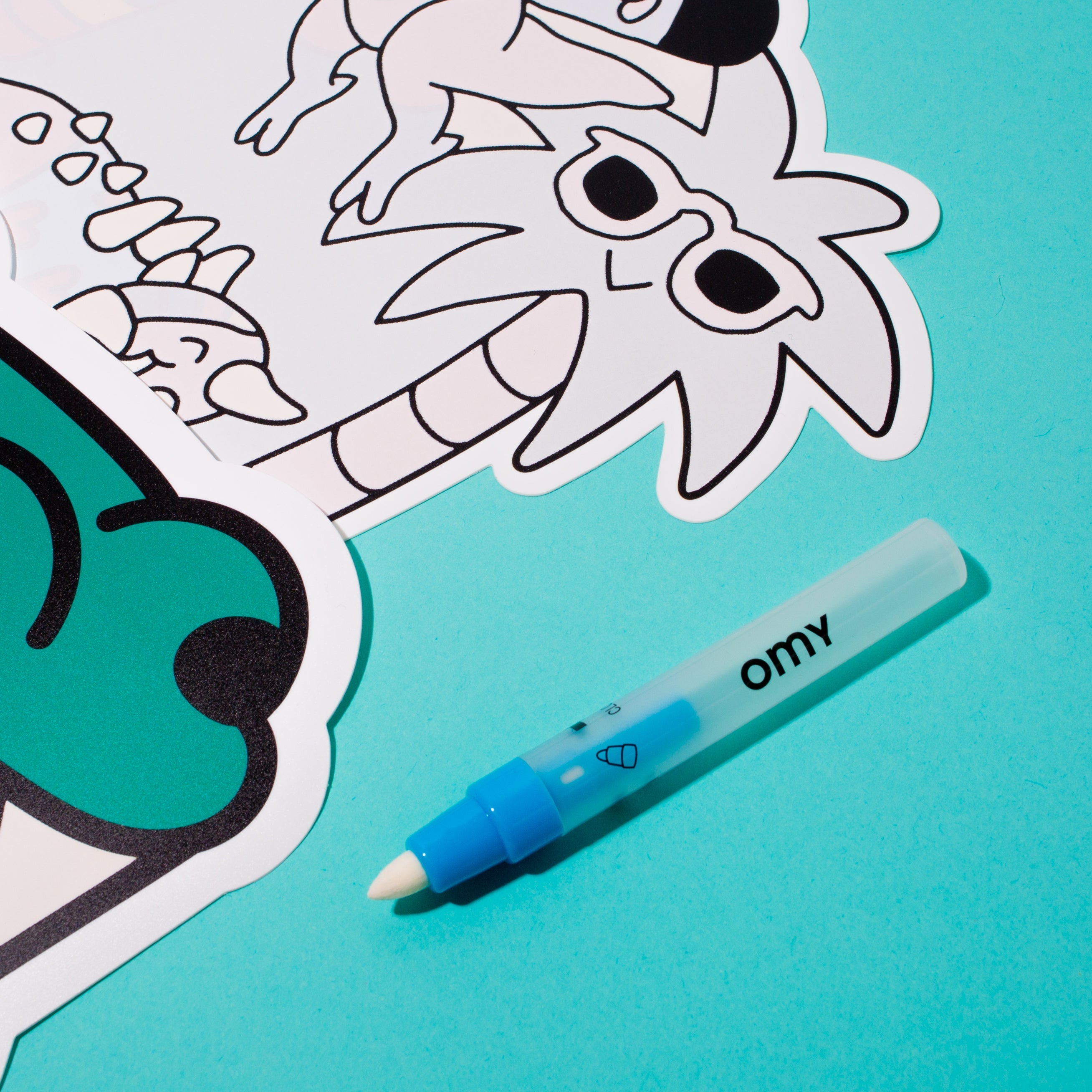 Gros plan sur le set de table Magic Set Dino OMY à colorier et son feutre bleu OMY, sur fond turquoise
