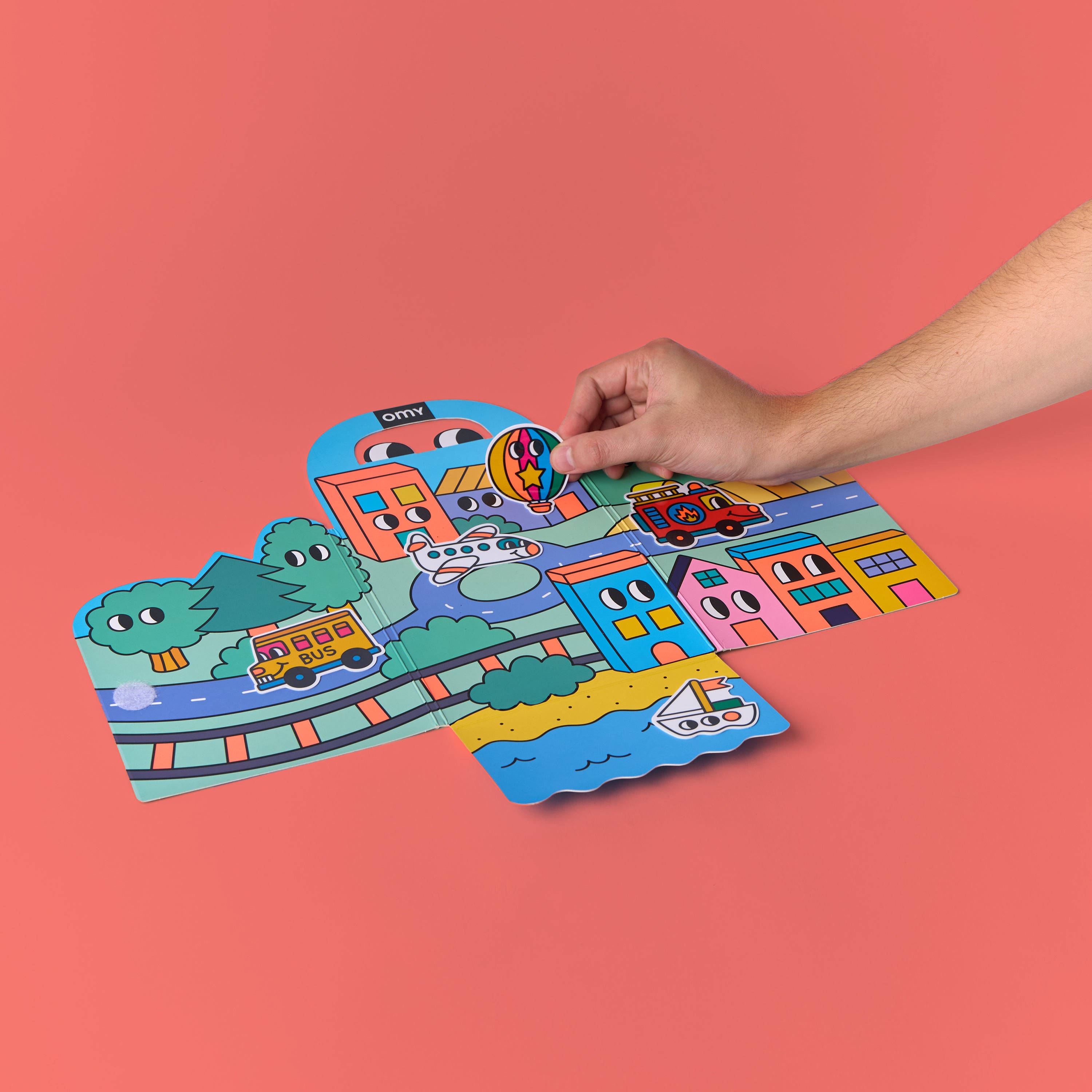 Kit d'autocollants Montessori - Vroom Vroom par OMY avec une main qui place un sticker sur une scène de ville colorée sur fond rose – activité interactive sans écran pour enfants