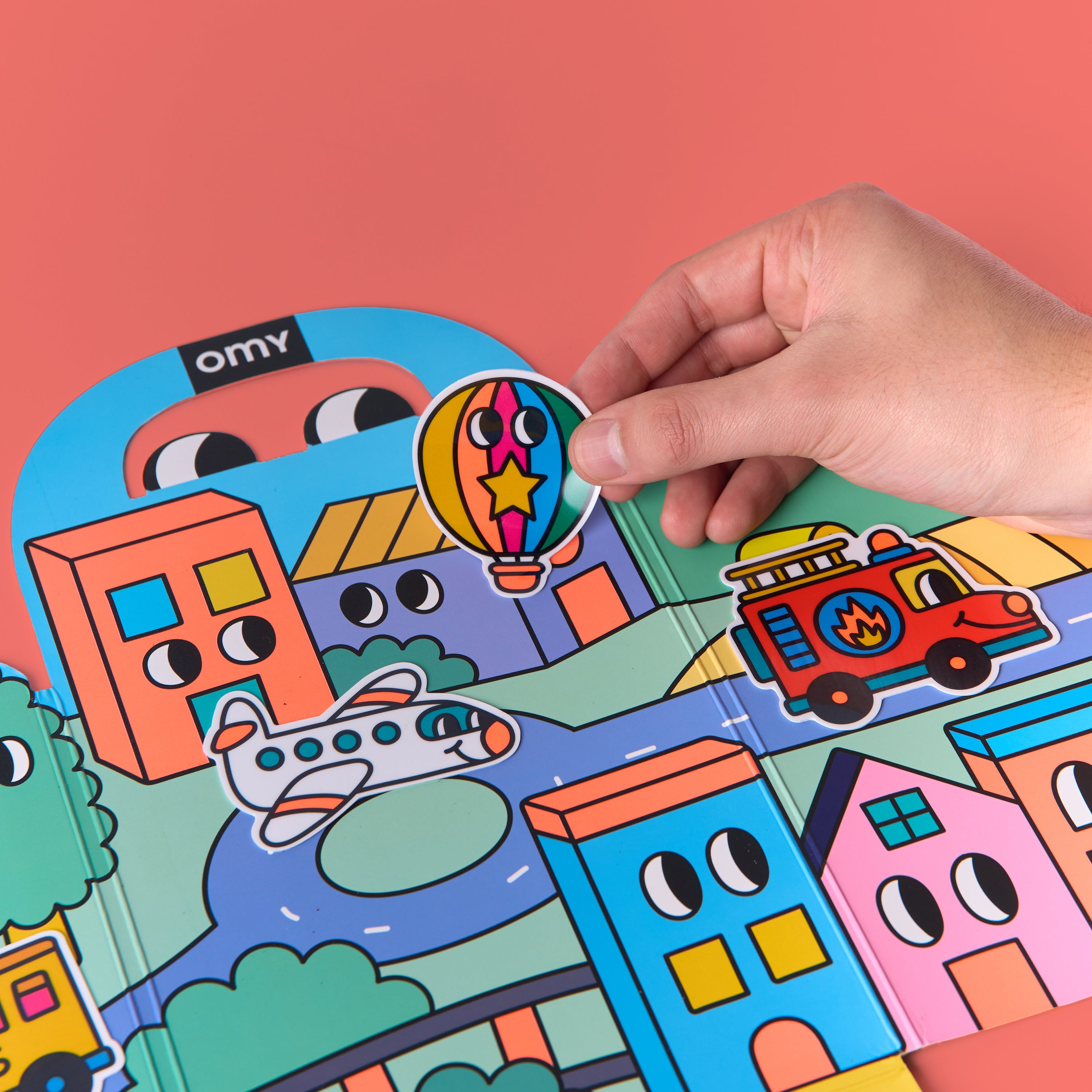 Main posant un sticker montgolfière sur le décor de ville coloré du Kit d'autocollants Montessori - Vroom Vroom par OMY avec avion et camion de pompiers sur fond rose – activité créative pour enfants