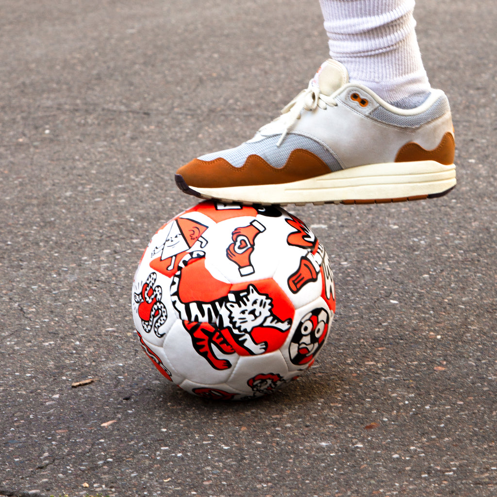 Fuß auf einem OMY-Fußball mit roten und schwarzen Motiven, darunter ein Tiger, auf dem Asphalt
