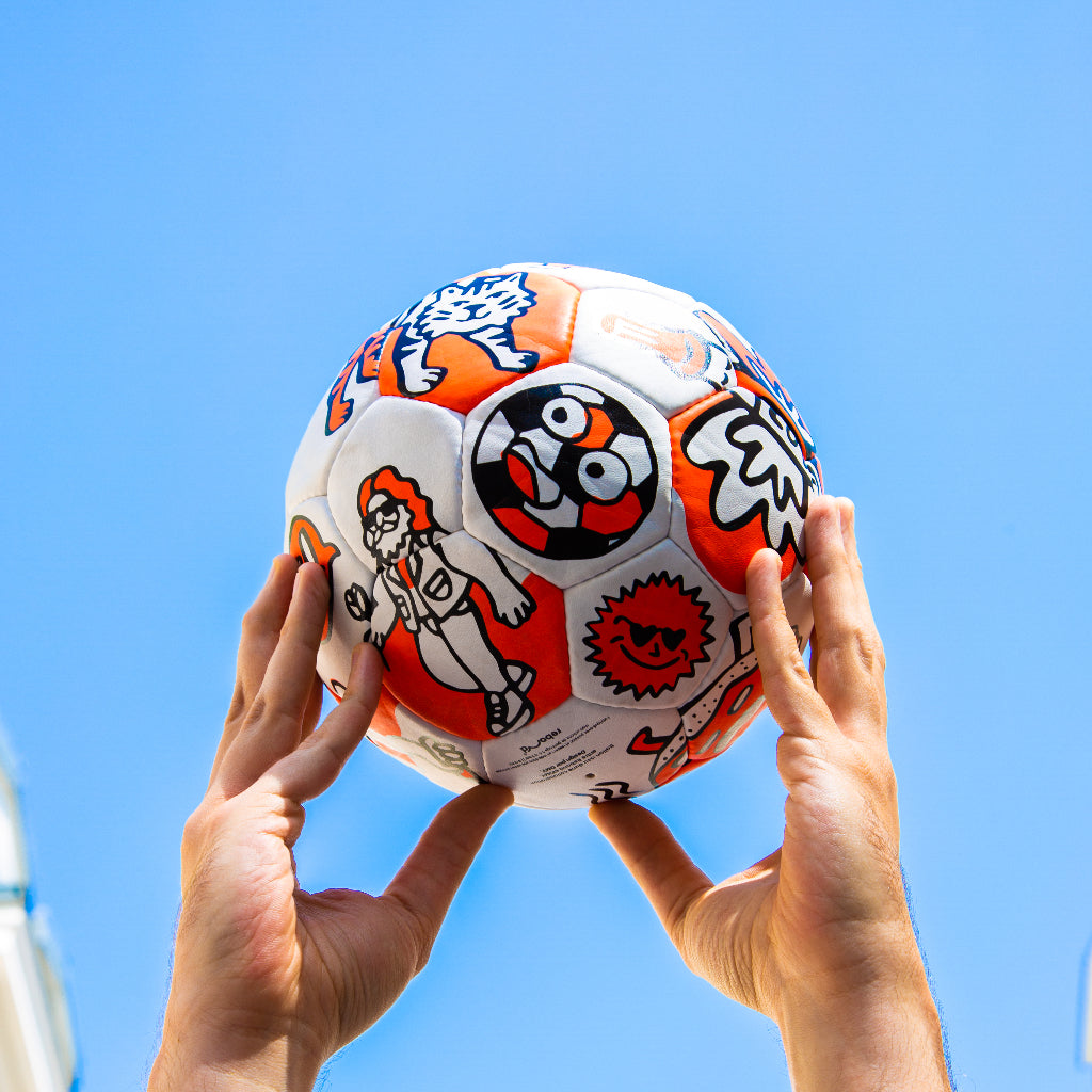Hände heben einen OMY-Fußball mit orangen und schwarzen Motiven gegen den blauen Himmel: Tiger, Sonne, lächelnder Ball