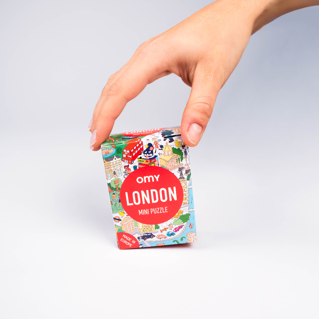 Hand houdt de doos van de OMY Mini Puzzle London vast, met monumenten en een rode Londense bus, op een lichtgrijze achtergrond