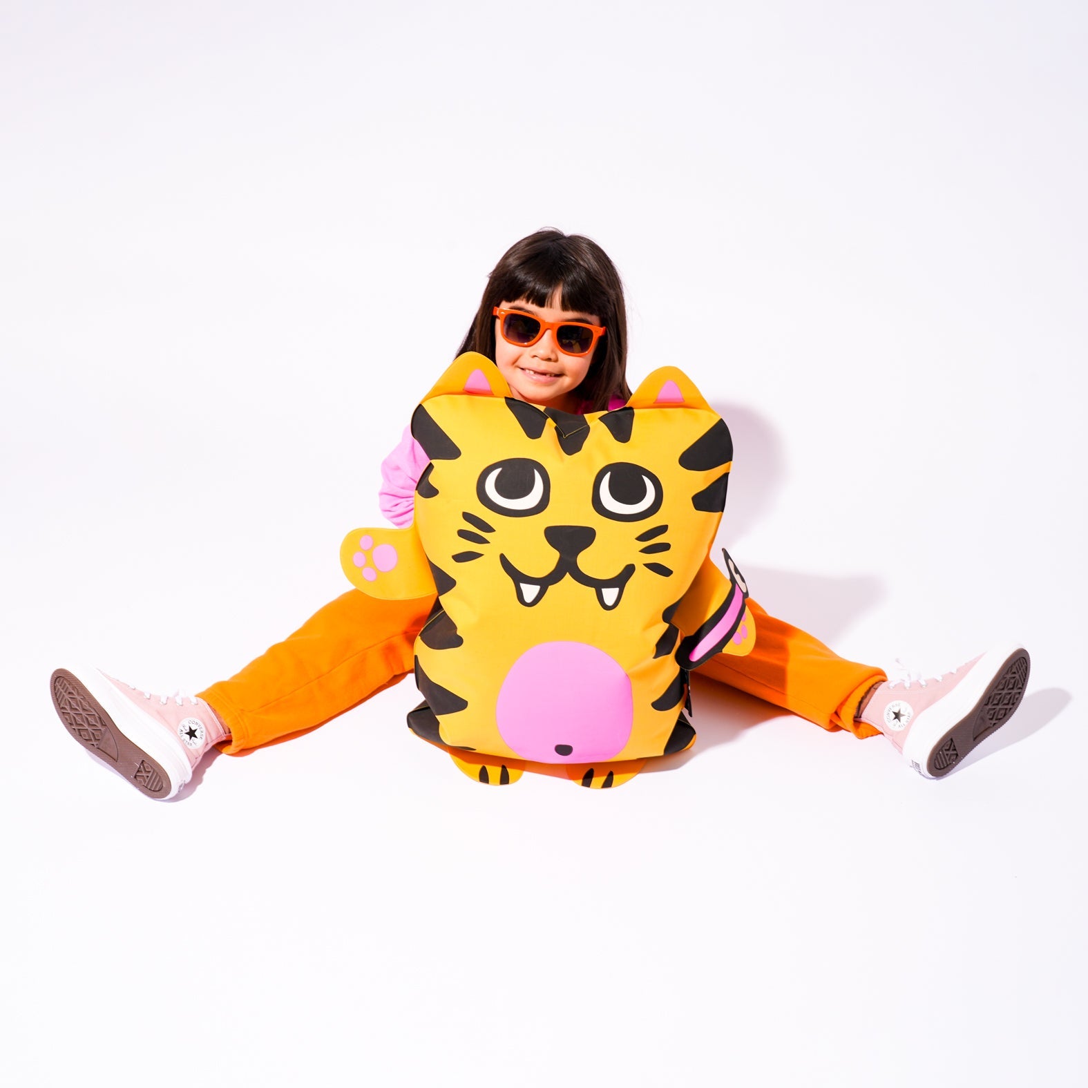 Fillette souriante à lunettes de soleil assise derrière le pouf gonflable OMY Tiga, tigre jaune rayé, sur fond blanc