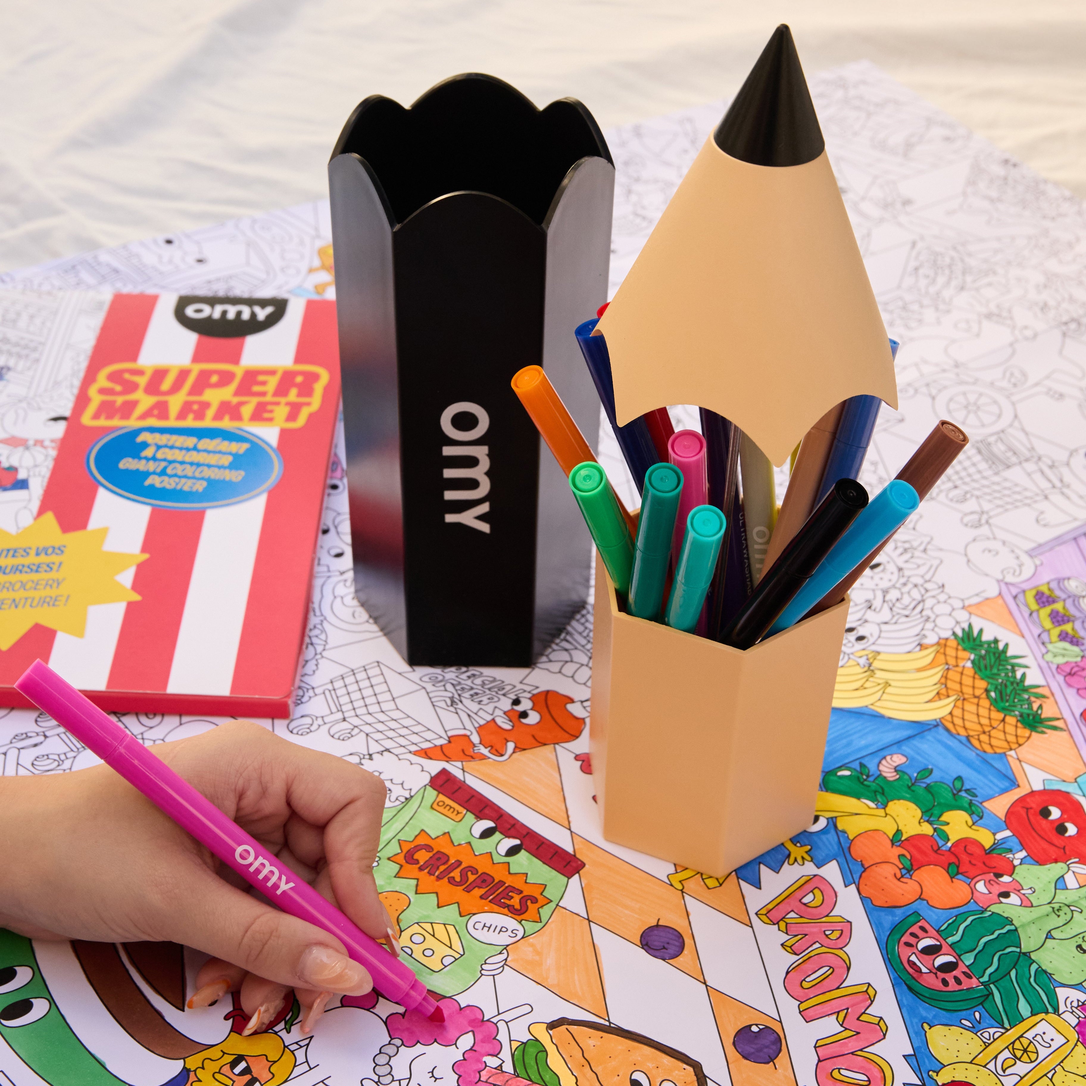 Main d’un enfant coloriant un poster géant OMY avec un feutre rose ; les deux boîtes à crayons et les feutres sont visibles sur la table.
