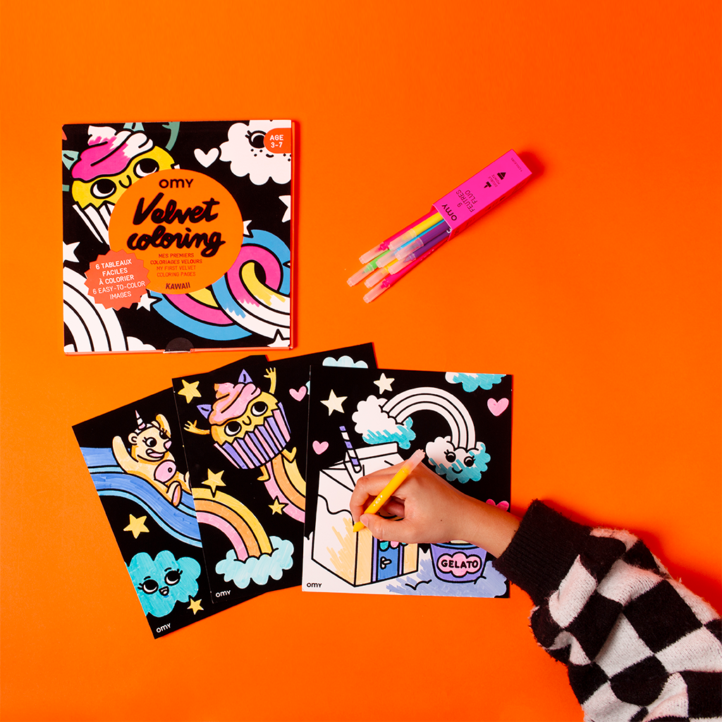 Confezione Velvet Coloring Kawaii OMY e una mano che colora con il pennarello i suoi quadri in velluto: unicorno, cupcake e cartone del latte, su sfondo arancione
