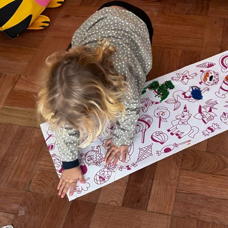 Enfant jouant au sol avec le rouleau adhésif Dreamland de OMY, illustré de dessins amusants à colorier comme des licornes, fruits et personnages rigolos.