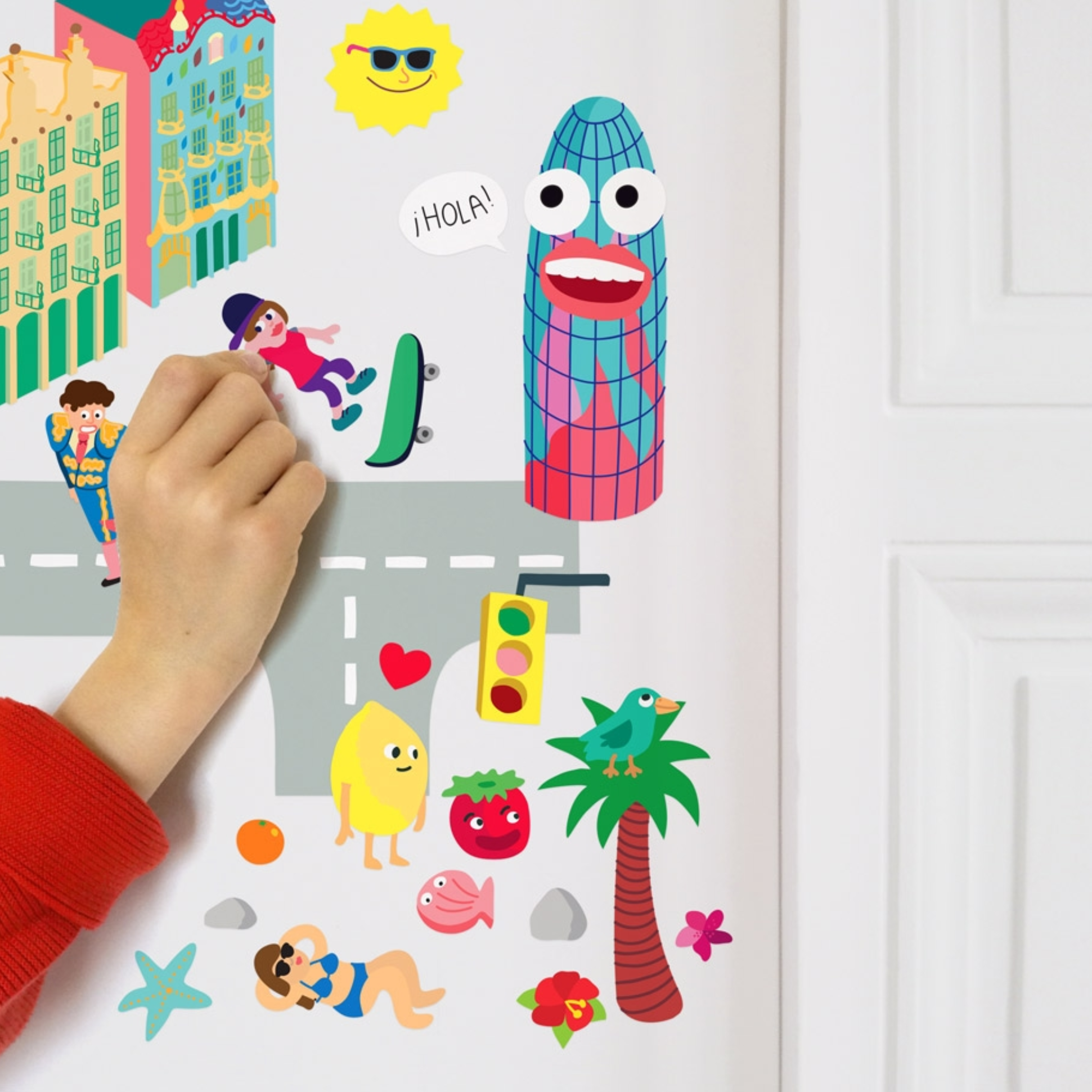 Main d'enfant collant les Wall Stickers Barcelona OMY sur un mur : tour Agbar, palmier, soleil et personnages