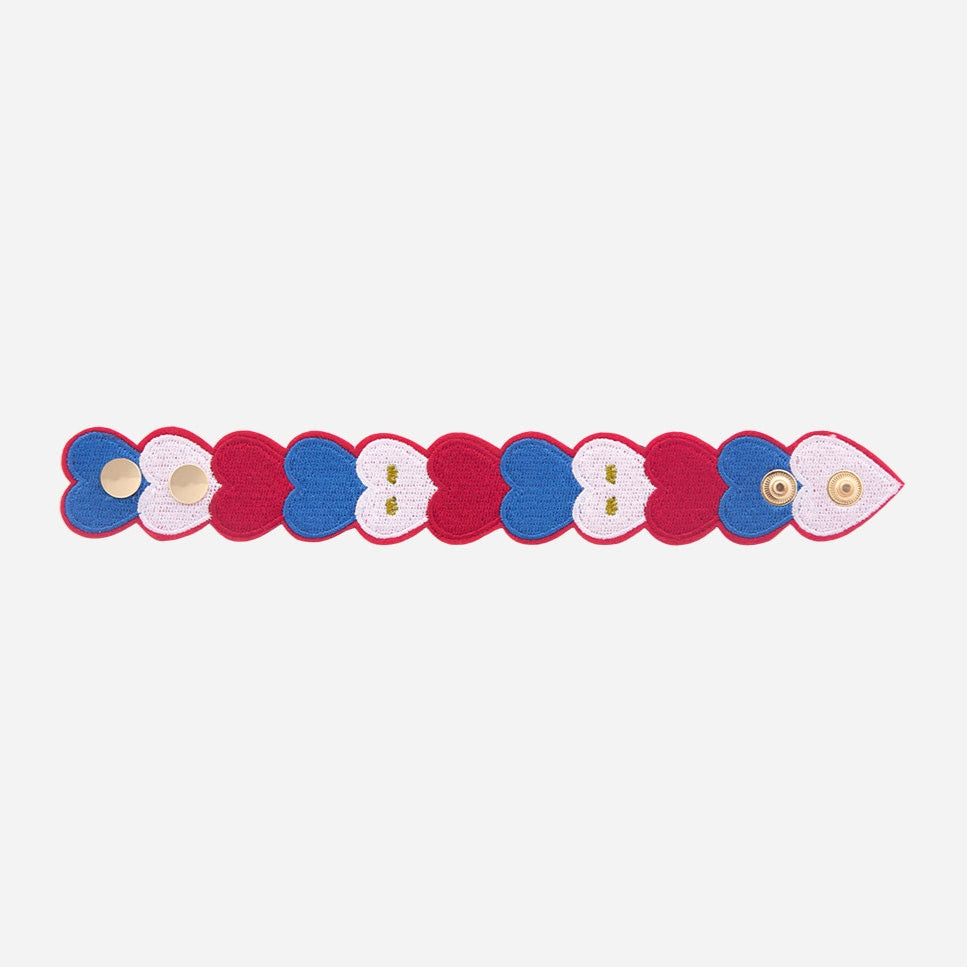 OMY geborduurde armband met blauwe, witte en rode hartjes, plat neergelegd met de gouden drukknopen, op een witte achtergrond