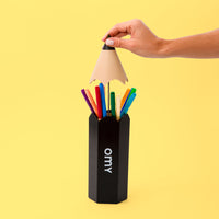 Hand hebt die Spitze der schwarzen Riesenstift-Box OMY an, die bunte Filzstifte freigibt, auf gelbem Hintergrund.