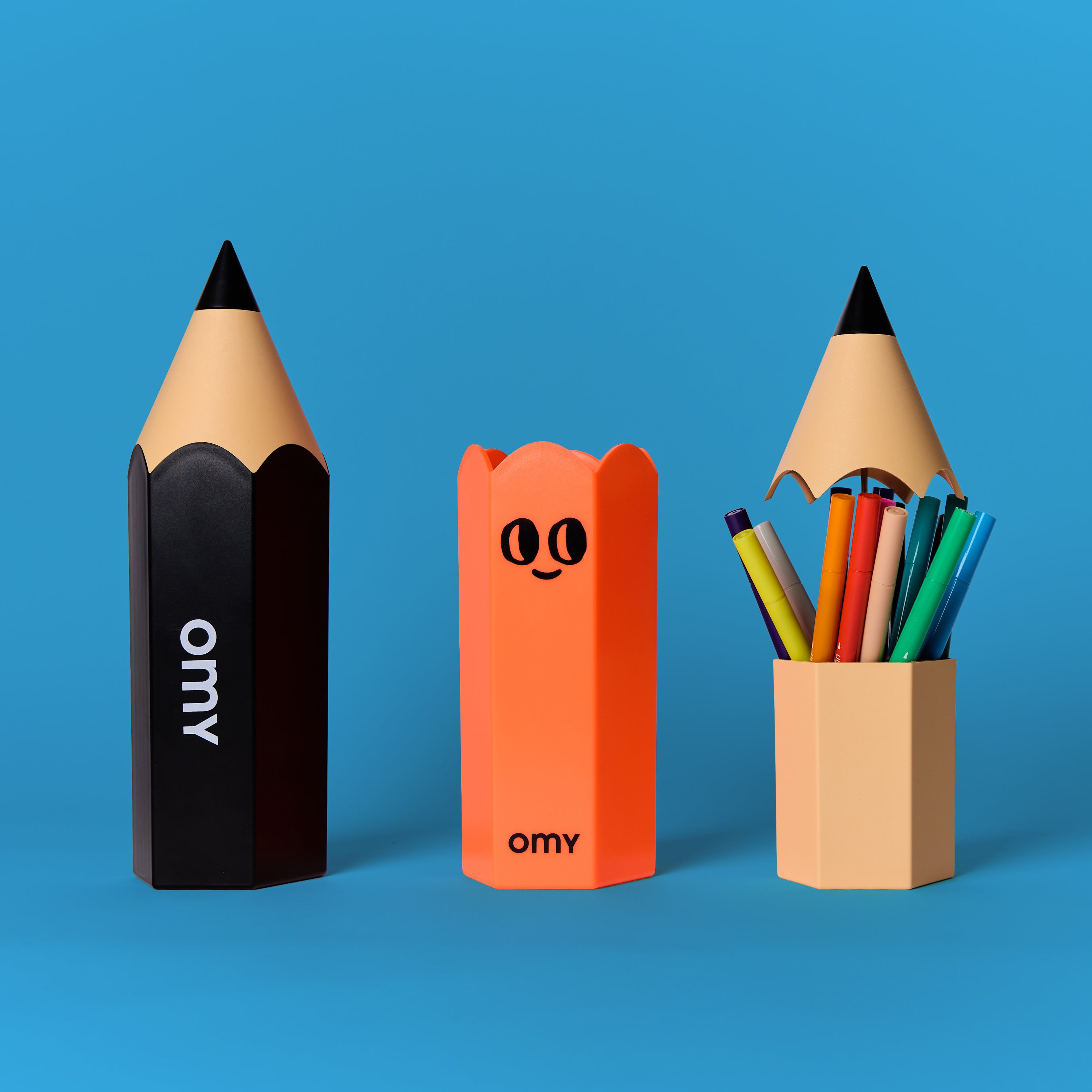 Trio de boîtes à crayons géantes OMY (noire, orange, beige) avec feutres colorés rangés dans l’une d’elles, sur un fond bleu éclatant.