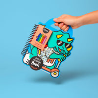 Hand holding the Dino OMY Super Coloring Book shaped like a triceratops with sunglasses on a scooter, coloring book with stickers and 5 wax crayons