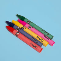 The 5 OMY smiley-face wax crayons included with the Dino coloring book: red, blue, yellow, pink and green, on a sky blue background