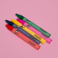 Los 5 crayones de cera OMY con cara sonriente incluidos con el cuaderno para colorear Lily: rojo, azul, amarillo, rosa y verde, sobre fondo rosa
