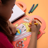 Little girl coloring a popcorn tub with a wax crayon in the Truck OMY coloring book, with sushi and pretzel stickers