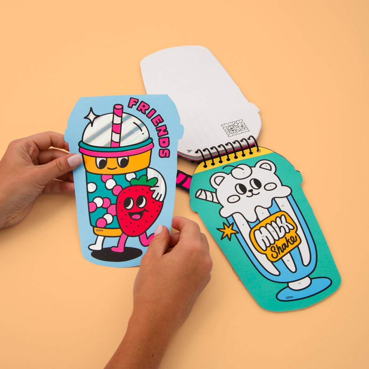 Mains tenant une carte Friends complétée en stickers du carnet Sticker by Number Bubble Tea OMY, ouvert sur Milkshake