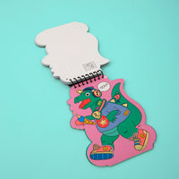 Dino OMY sticker activity book open to a green dinosaur decorated with stickers, pink page, on a turquoise background.