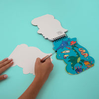 Hands peeling a sticker off the Dino OMY sticker activity book open to an underwater scene, on a turquoise background.