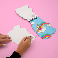 Carnet d'activités stickers Lily OMY ouvert sur un décor de licorne et d'arcs-en-ciel, avec une main écrivant sur une carte postale détachable
