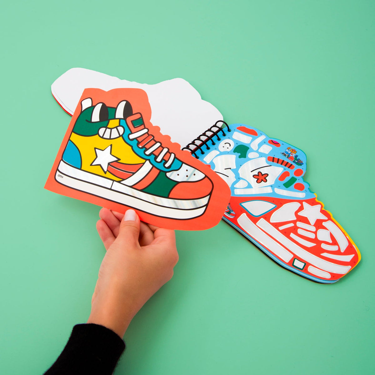 Street OMY sticker book shaped like a sneaker, held in hand, pages of colorful stickers open, on a mint green background.