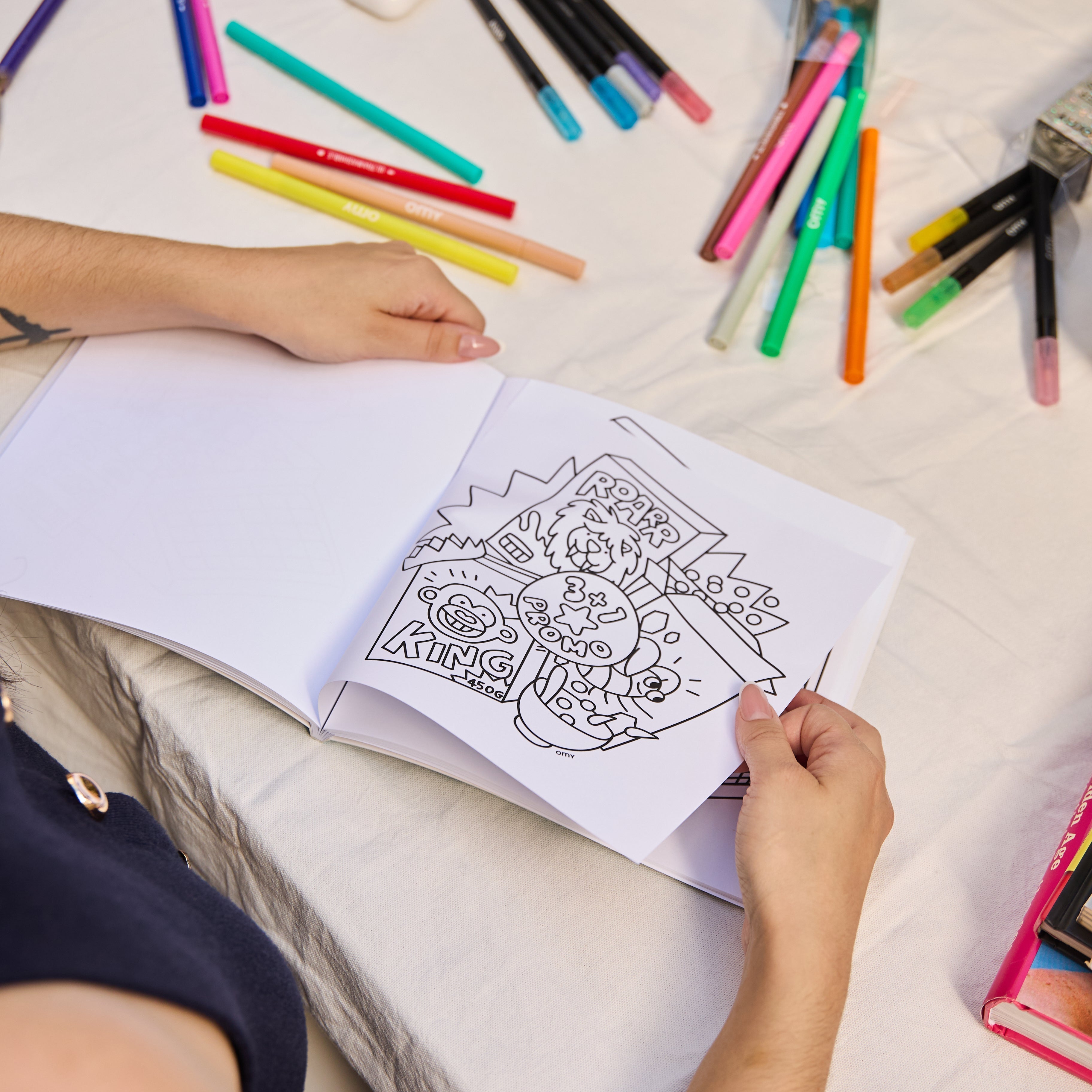 Cozy Art Super Market coloring book open on a table, child's hands holding an illustrated page with comic-style drawings, surrounded by OMY colored markers
