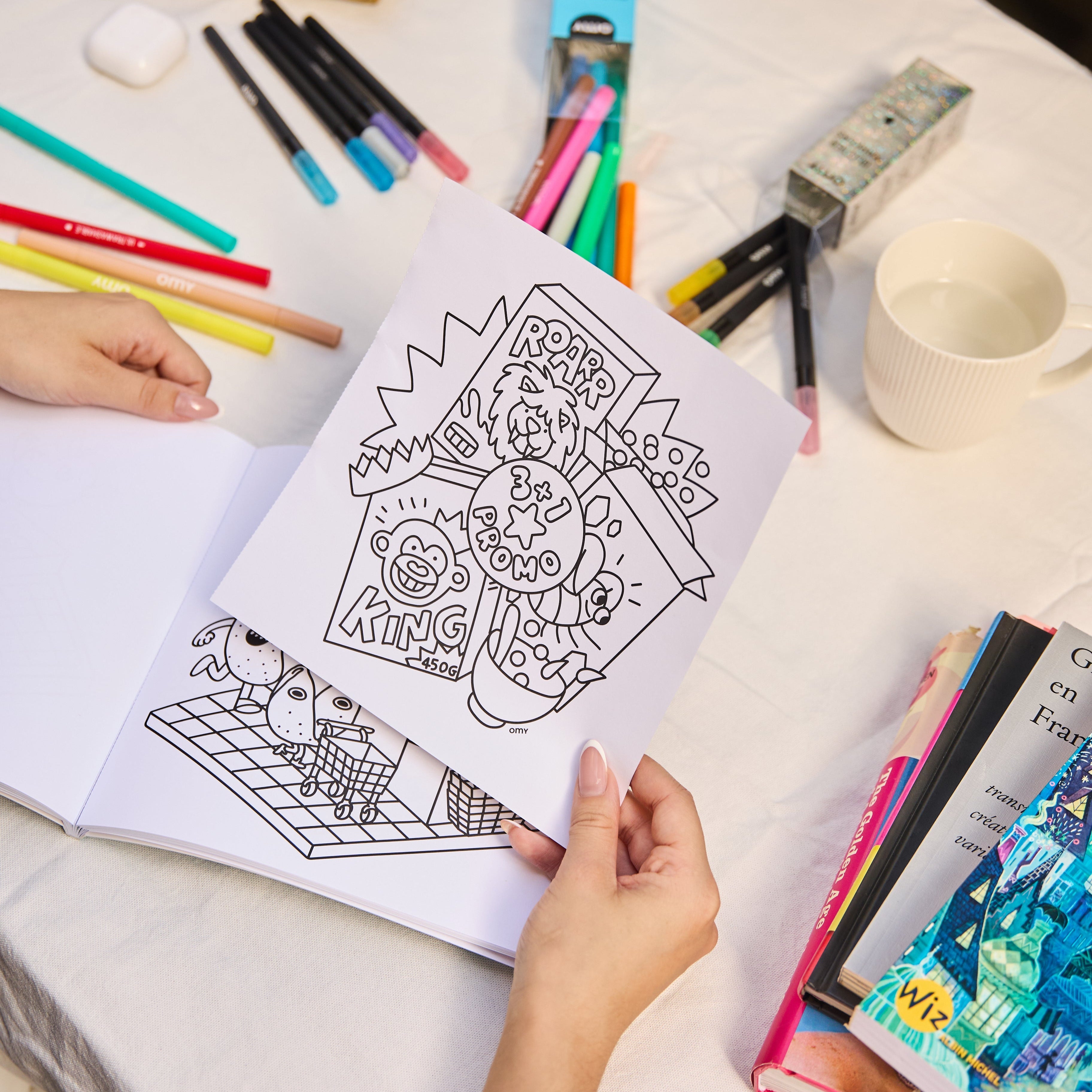 Page from the Cozy Art Super Market notebook with an illustration to color of food products, held by a hand above the open notebook on a table lined with markers and books