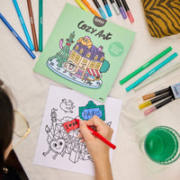 Kind kleurt een “PARIS”-illustratie uit het Cozy Art notitieboek van OMY met een rode stift, notitieboek en stiften zichtbaar op de achtergrond
