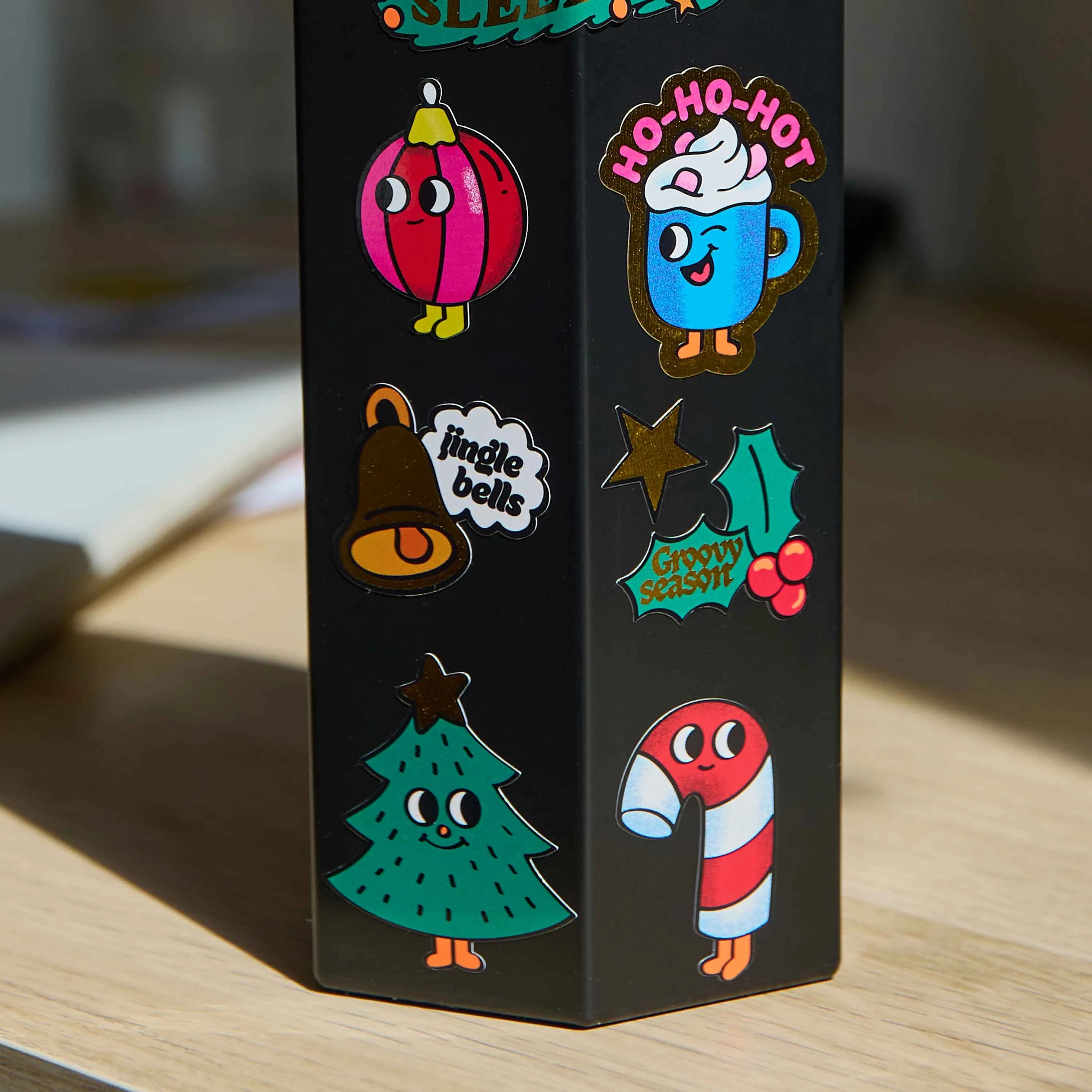 Stickers navideños grandes OMY aplicados sobre un objeto negro, con abeto, bola de Navidad, taza, campana, acebo y bastón de caramelo.