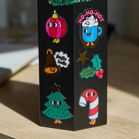 Stickers navideños grandes OMY aplicados sobre un objeto negro, con abeto, bola de Navidad, taza, campana, acebo y bastón de caramelo.