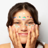 Enfant souriante portant les stickers visage Kawai OMY : arc-en-ciel, étoiles filantes et petits motifs sur le front et les joues