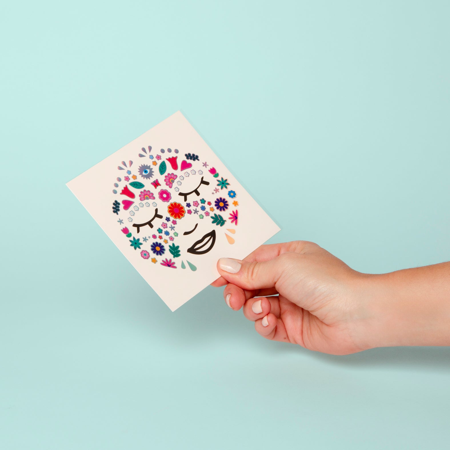 Planche de Face Stickers Flower OMY tenue en main : fleurs et feuillages colorés dessinant un visage, sur fond vert d'eau