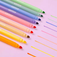 Les 9 feutres pastel double pointe OMY alignés en dégradé de couleurs, avec un trait de chaque teinte sur fond rose