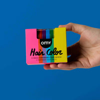 Main tenant la boîte Hair Color OMY arc-en-ciel : 6 craies temporaires pour cheveux, sur fond bleu
