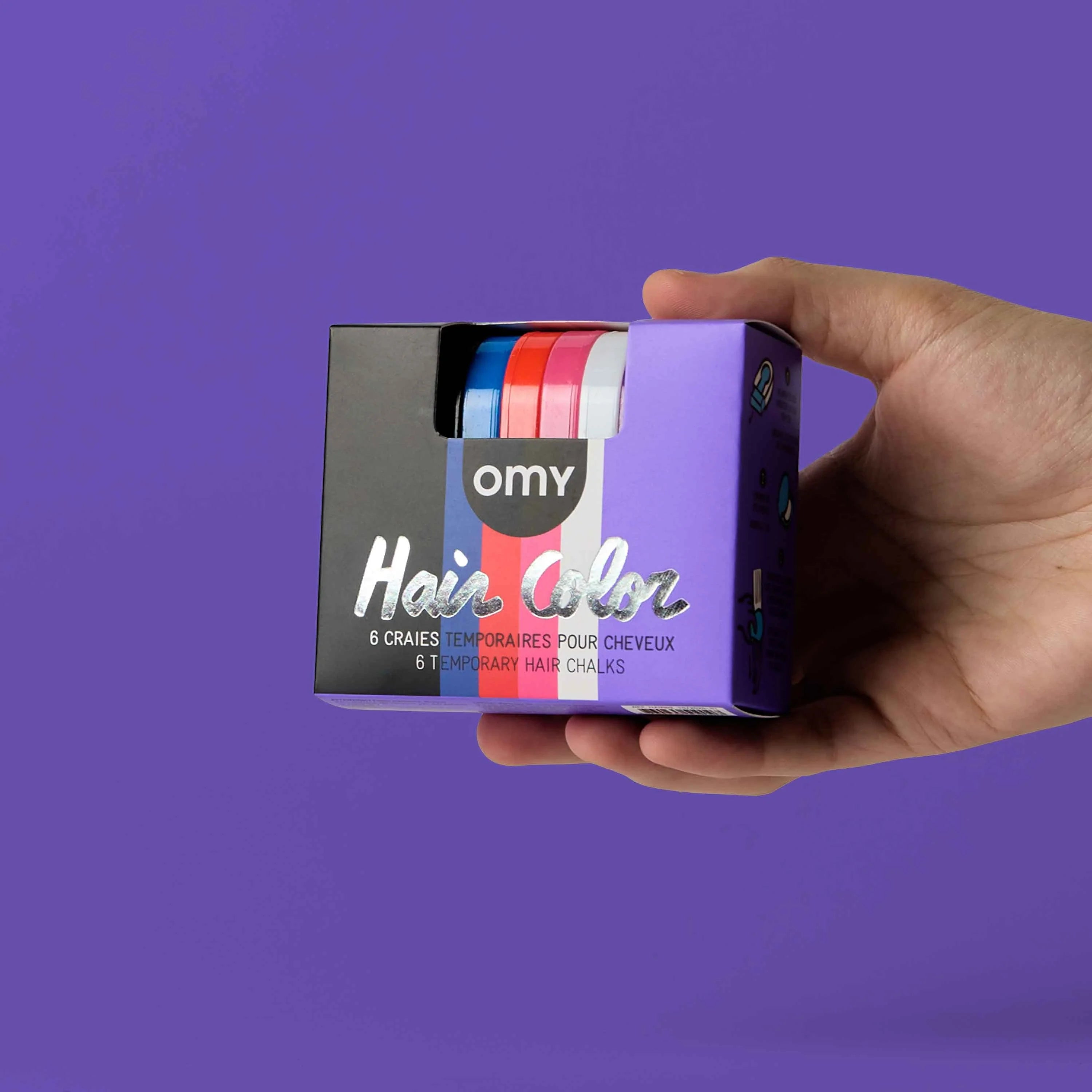 Main tenant le coffret Hair Color Wicked OMY de 6 craies temporaires pour cheveux, sur fond violet