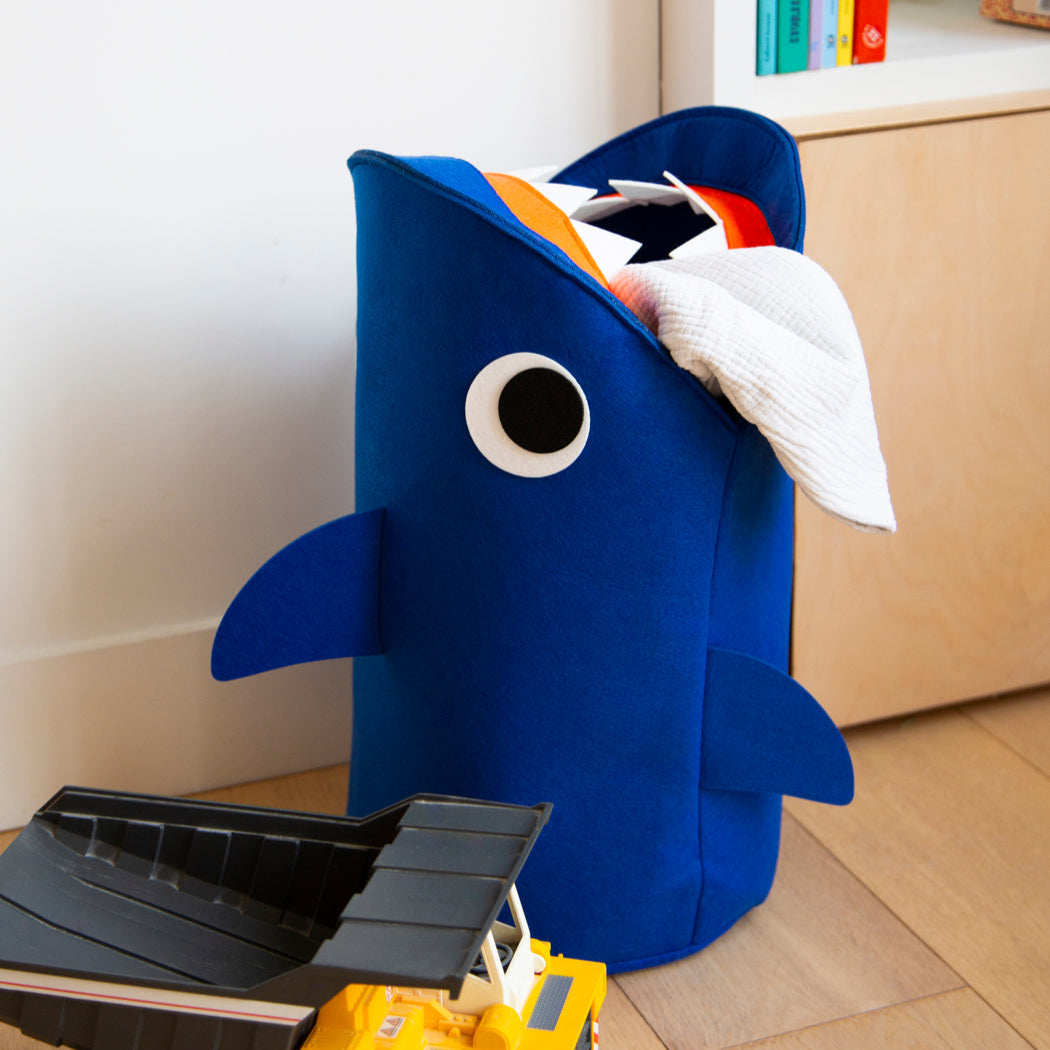Tidy Bag Sharky OMY, blauer Aufbewahrungskorb aus Filz in Form eines Hais, gefüllt mit Wäsche, neben einem Spielzeuglaster.