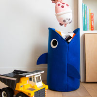 Tidy Bag Sharky OMY, blauer Aufbewahrungskorb für Kinder aus Filz in Form eines Hais, mit Kuscheltier und Laster in einem Kinderzimmer.