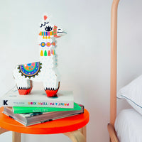 Lamp 3D Lama OMY en carton, lama blanc aux motifs colorés, posée sur des livres et un tabouret orange dans une chambre