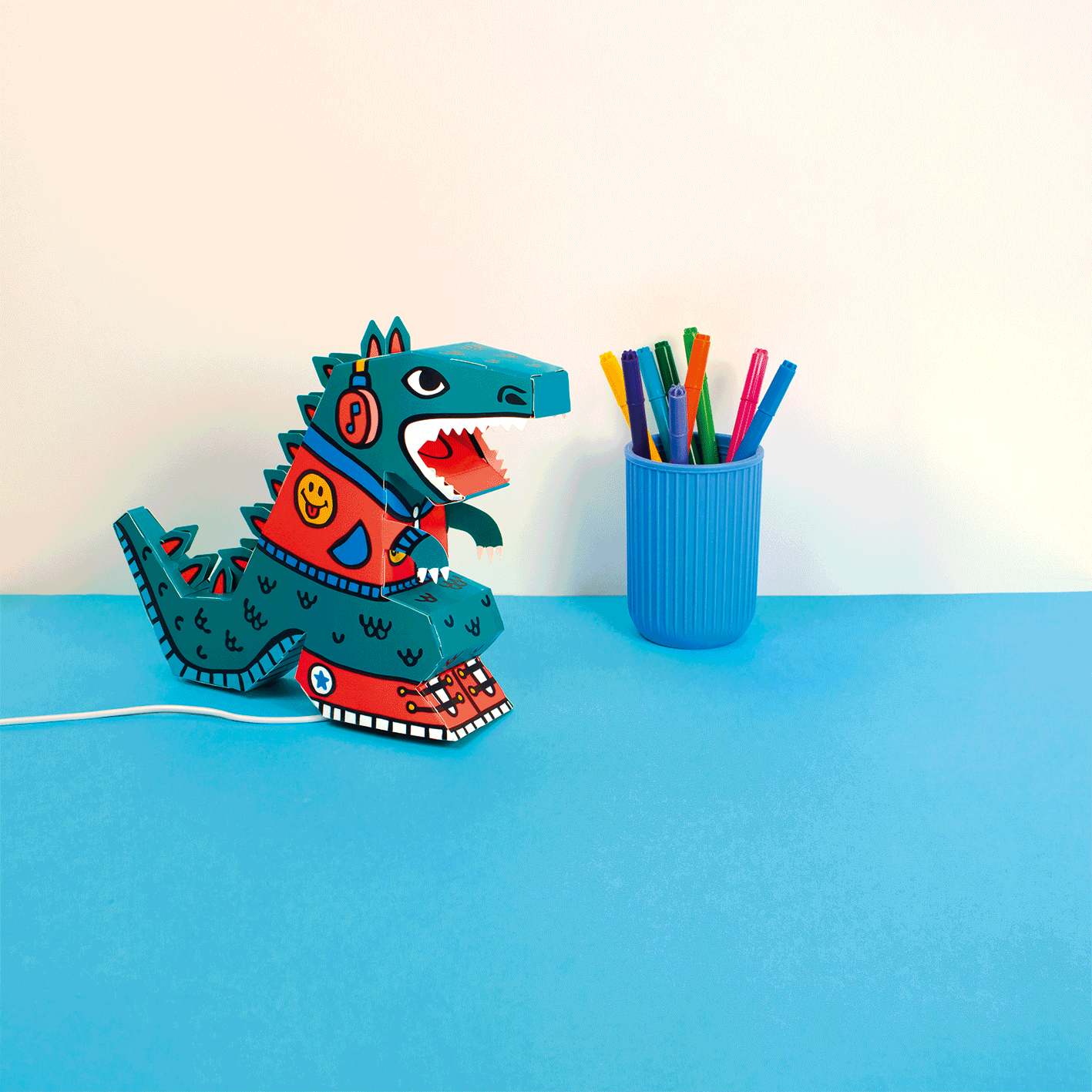 Animation der OMY Dino 3D-Lampe aus Karton auf einem blauen Schreibtisch, neben einem Stiftebecher mit Filzstiften.