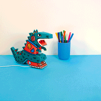 Animation de la Lamp 3D Dino OMY en carton montée sur un bureau bleu, à côté d'un pot de feutres